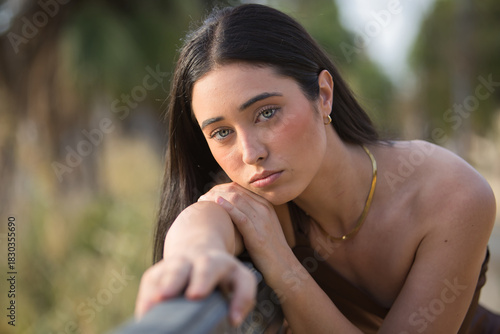 Portrait of a young, beautiful, dark-haired, blue-eyed Hispanic woman wearing a brown chiffon dress, with a sweet and tender gaze, her face resting on her arm and leaning on a metal railing.