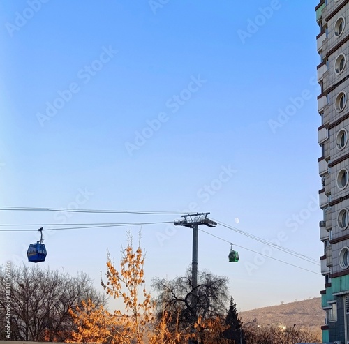 Cable cars traveling above the city at sunset time, with bare trees, residential buildings and a visible moon in the clear sky. 