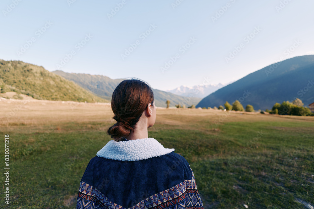 Obraz premium Woman back view looking at expansive mountain valley landscape in nature outdoors, contemplative travel scene with distant peaks, grassy meadow and clear sky at golden hour.