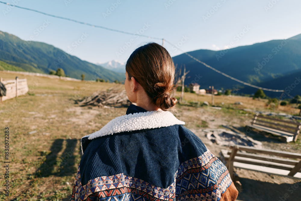 Naklejka premium Woman back view in rural countryside wearing patterned jacket, overlooking mountain landscape and valley. Outdoor travel scene with natural light, calm morning and wooden fence.