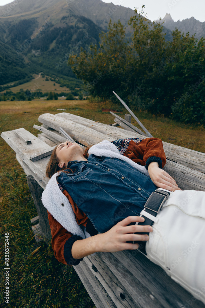 Naklejka premium woman bench mountains nature relaxed jacket denim outdoors lying on a wooden seat in a scenic meadow, casual fashion portrait showing leisure, travel and peaceful rest in countryside autumn