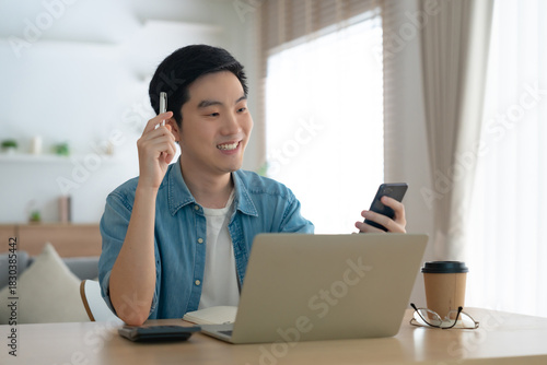 Happy young Asian man working on laptop in living room. Freelance worker male sitting at desk writing notes while watching webinar, studying online