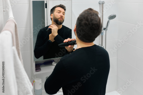 Man grooming beard in bathroom mirror while using electric trimmer during morning routine