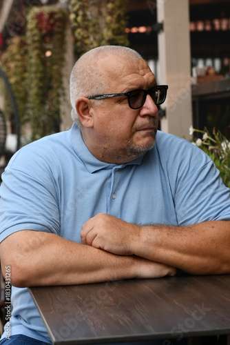 A bearded older man wearing sunglasses and a blue T-shirt sits at a table in a street cafe.