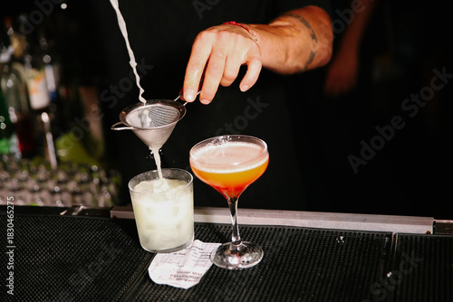 Professional mixologist pouring and straining a craft cocktail from a metal shaker into a row of coupe glasses on a dark bar counter