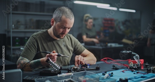 Adult Male Worker Closely Soldering Small Drone Component on Workbench, With Colleagues Working in Blurred Background. Concept of Collaborative Environment of Technology Production Facility.
