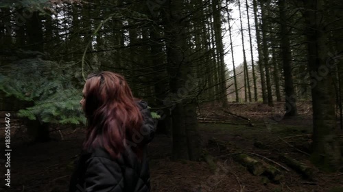 A profile close-up shot of a woman in a dark hooded jacket looking upwards at the dense, dark green branches of a spruce or fir tree in a forest. The scene evokes nature and contemplation.