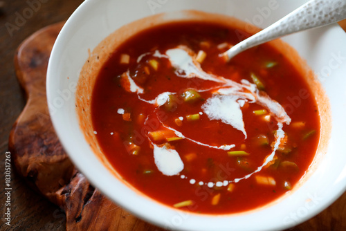 COLOURFUL TOMATO BASED VEGETABLE SOUP WITH STYLISH SWIRL OF PLANT BASED CREAM IN WHITE BOWL WITH BEATON METAL SPOON ON RUSTIC WOODEN BOARD .