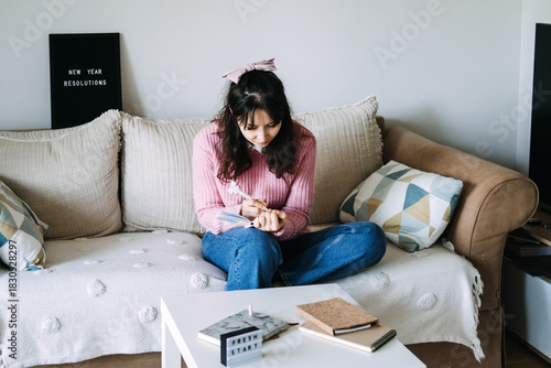 Young adult woman sits on sofa writing goals in small notebook next to letter board displaying NEW YEAR RESOLUTIONS. Financial goals, money mindset, budgeting goals, financial literacy.