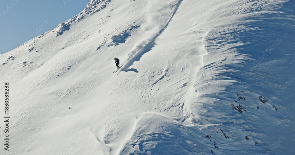 Fototapeta premium Aerial photo of a free ride skier carving through fresh powder snow on a steep alpine slope. Drone shot capturing the thrill of first tracks in pristine winter mountains.