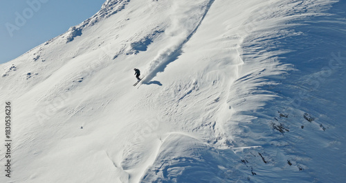 Aerial photo of a free ride skier carving through fresh powder snow on a steep alpine slope. Drone shot capturing the thrill of first tracks in pristine winter mountains.