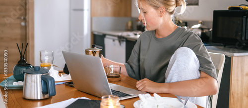 Young woman working remotely at home kitchen table with laptop, coffee, and breakfast, representing freelance lifestyle, flexible schedule, and modern work-from-home concept.