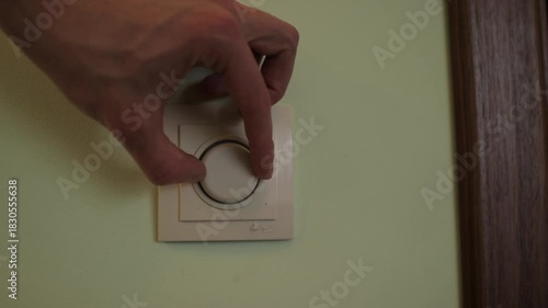 Male hand turning a round knob on a modern wall dimmer switch. Adjusting the brightness of the light and saving energy at home or office
