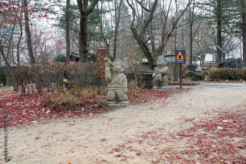 Stone statues stand along an autumn pathway covered in red leaves, surrounded by bare trees and rustic scenery, creating a peaceful cultural atmosphere in the forest park.