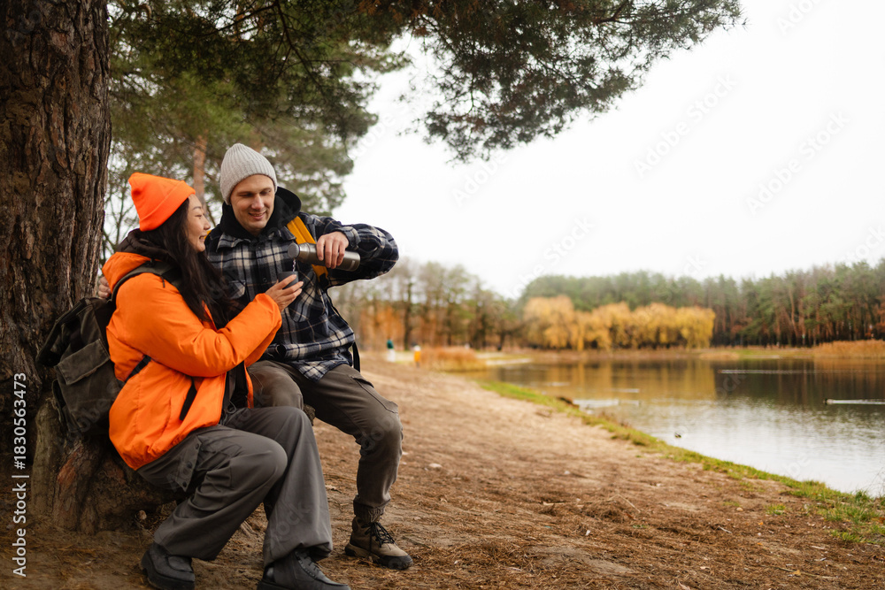 Fototapeta premium Hiking couple enjoys nature taking a break with water bottle or hot drink tea near lake in a lush forest during a cold day.