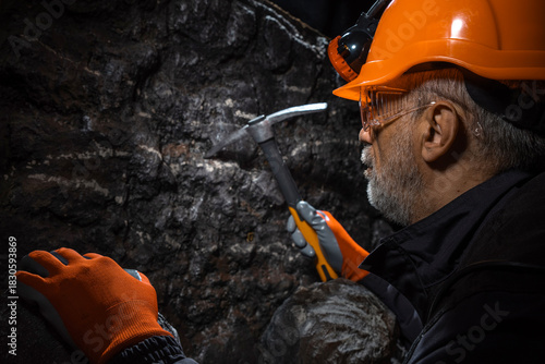 A man in a hard hat with a flashlight examines rock using a pickaxe. Geology, speleology, mining with a scientific approach