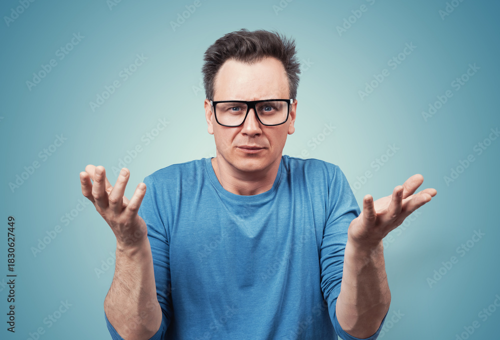Naklejka premium Confused man in glasses raising his hands in a questioning gesture, expressing frustration or disbelief. Casual blue t-shirt, puzzled facial expression. On blue background.