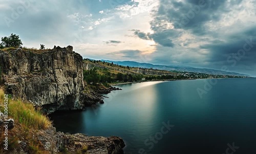 Dramatic cliffside view of Okanagan Lake, with cloudy skies and reflections enhancing the serene water surface during sunset