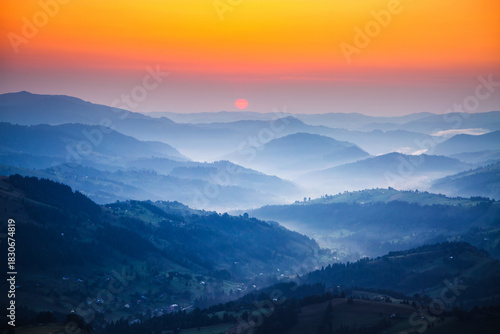 Breathtaking view from above captures the sunset behind layers of mountains surrounded by soft mist in the valleys, creating a calm atmosphere at dusk. Photo wallpaper. Discovery the beauty of earth.
