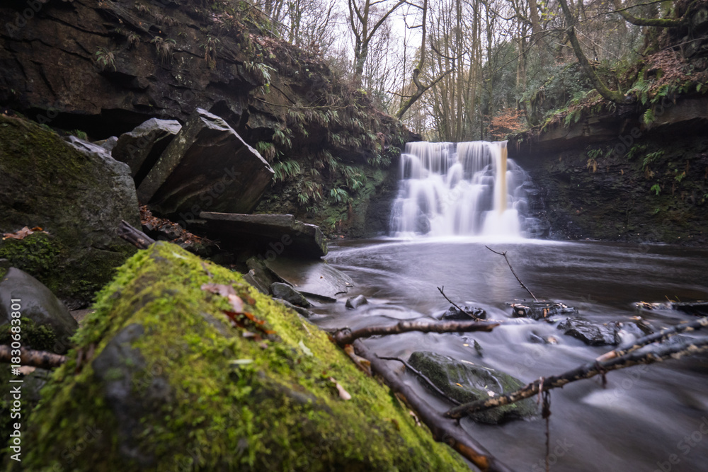 Obraz premium Long-exposure of a tranquil woodland waterfall on an autumn day. Moss and leaf covered rocks, soft flowing water, and rich forest textures create a peaceful natural landscape ideal for outdoor themes.