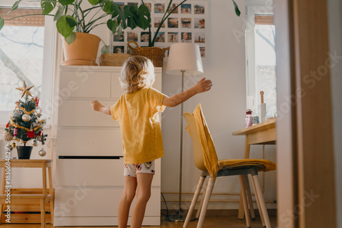 Playful child jumps and dances in messy living room decorated with small Christmas tree. Happy family moments. Funny boy playing at home at warm sunny winter morning.