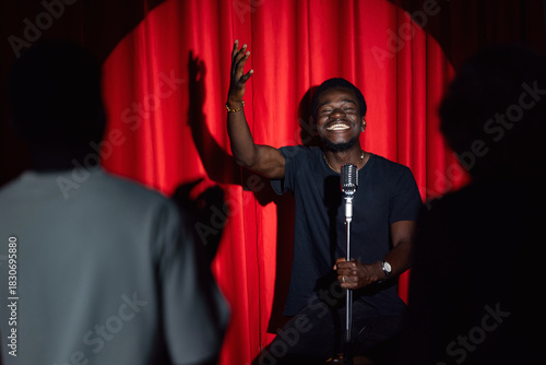 Tableau sur toile Happy young african american man comic tells stand up monologue on stage with red curtains