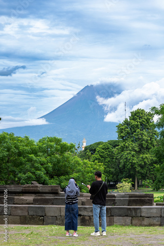 Java Indonesia 12 11 2025 – Two local tourists taking photos from behind with Mount Merapi rising in the background on a clear day.