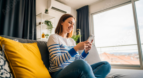 Smiling woman sitting on couch using smart phone in modern apartment home