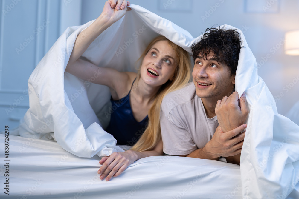 Obraz premium Happy couple under blanket smiling and looking up with excitement in bed. Concept of love, intimacy, bedtime fun, romantic relationship, joyful morning, couple bonding moment.