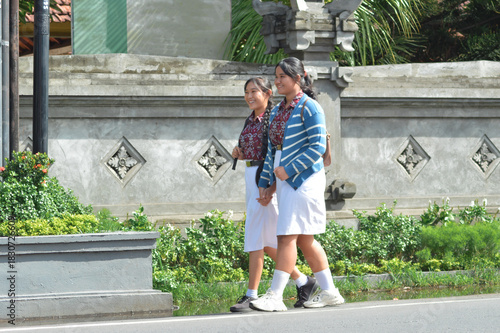 Two cheerful students in patterned uniforms, one with a striped sweater, walk side by side holding hands and chatting as they pass a decorative stone wall on their way to school.