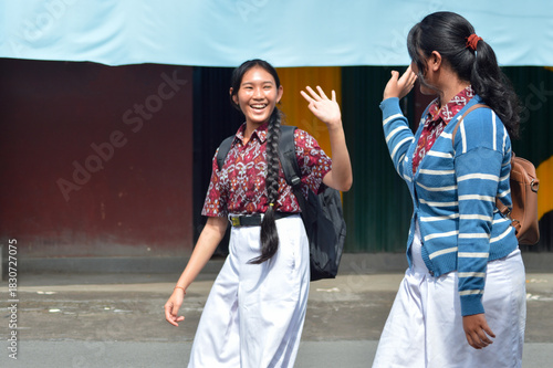 Two cheerful students in patterned uniforms with school bags walk along a street, smiling and waving to each other as they part ways at a junction, heading home after classes.