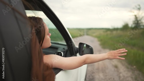 Passenger stretches arm and hand out from car window over countryside road with grass field and side mirror visible along empty asphalt path under pale cloudy sky green verge distant tree line