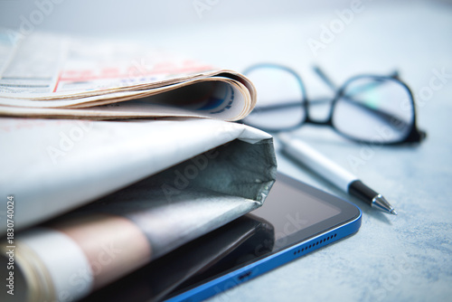 A pile of newspapers ,glasses and digital tablet on the table.