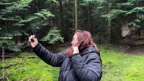 A side-profile shot of a female blogger or hiker in a dark jacket holding an action camera up to film herself against a dark, dense pine forest background. 