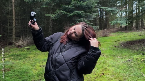 A side-profile shot of a female blogger or hiker in a dark jacket holding an action camera up to film herself against a dark, dense pine forest background. 