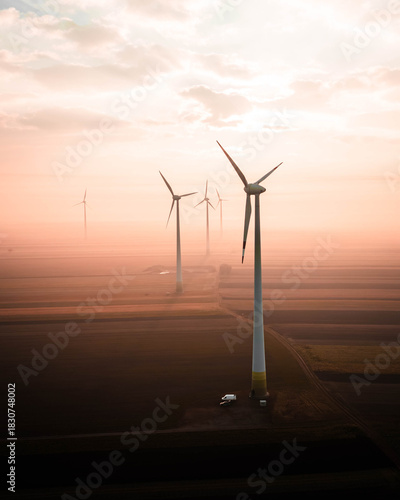 Wind turbines rising above soft morning fog, illuminated by warm golden sunrise tones. The calm landscape blends mist, light and motion, creating a serene and atmospheric view of modern renewable ener