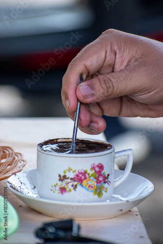 Hand stirring a traditional coffee drink in a decorative floral cup and saucer on a wooden table. Warm atmosphere, fresh brew, everyday lifestyle and caffeine routine scene.