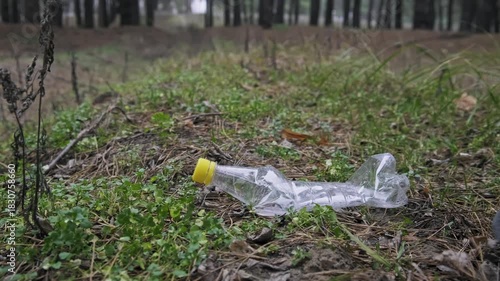 Legs in black sneakers walk past a discarded plastic bottle on the forest ground. This low-angle footage highlights human negligence and the issue of litter in nature.