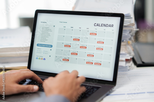 A hand checks a laptop calendar to manage a deadline timeline with an overworked task planner in the office.
