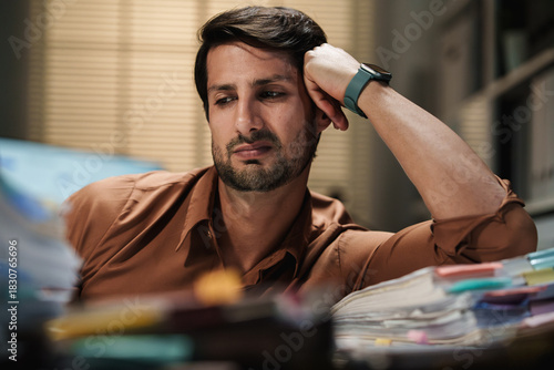 A bored, unhappy man sits exhausted in the office at night, feeling overworked, distressed, and sad.