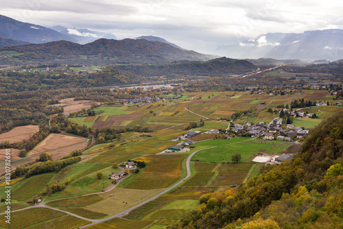 Scenic aerial panorama view of French alps in Savoie region, rocky mountain with vineyard and village, cloudy drama sky during autumn, with golden color on nature.