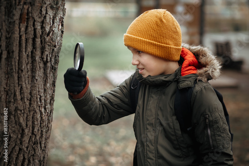 A curious child in a yellow beanie and green winter jacket examines a tree trunk with a magnifying glass in a forest.