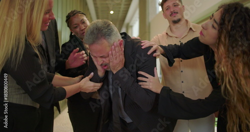 Distressed male professional showing anguish and worry while being comforted by supportive colleagues in office hallway during emotional workplace moment