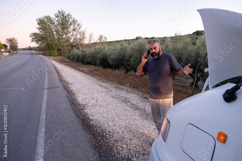 A middle-aged bearded man stands next to his camper van with the hood open, talking on the phone on a rural road lined with trees. The evening light enhances the tense situation.