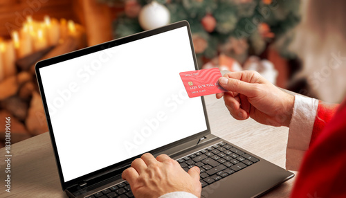 Santa holding credit card while using laptop with blank screen in warm Christmas interior, ideal for holiday mockups, ecommerce concepts and festive digital marketing
