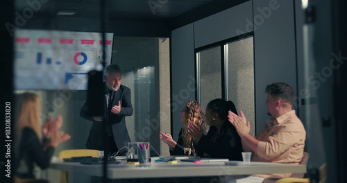 Diverse team applauding business leader following successful data presentation in modern corporate boardroom with screen displaying charts