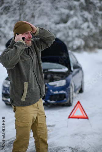 Man standing by broken car in snow, making a phone call for help