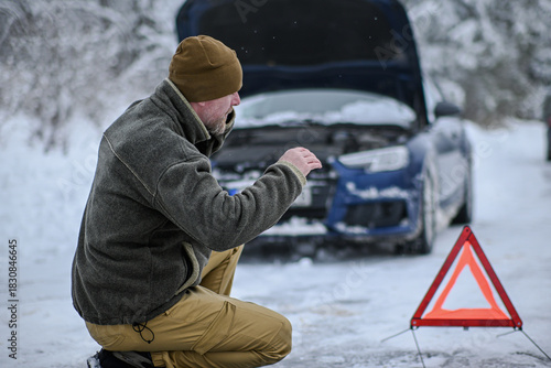 Man making a phone call, standing next to a broken car and warning triangle in snow