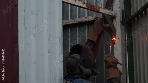 A welder works on a metal gate, creating bright sparks as he welds the steel surface.