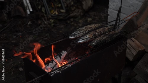 Three mackerel are grilled over an open fire, forming a golden-brown crust. The fish rest on the grill, enveloped in smoke.